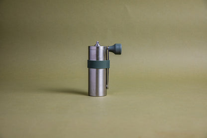 Stainless steel cylindrical shaped coffee mill canister with forest green silicone band holding stainless steel handle with plastic knob. Set against an earth-tone background.
