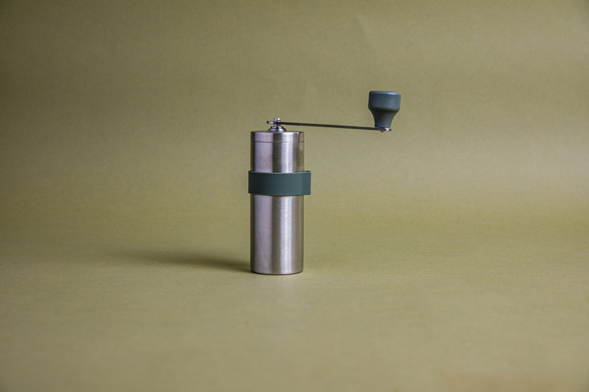 Stainless steel cylindrical shaped coffee mill canister with forest green silicone band stainless steel handle with plastic knob. Set against an earth-tone background.