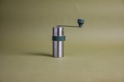 Stainless steel cylindrical shaped coffee mill canister with forest green silicone band stainless steel handle with plastic knob. Set against an earth-tone background.