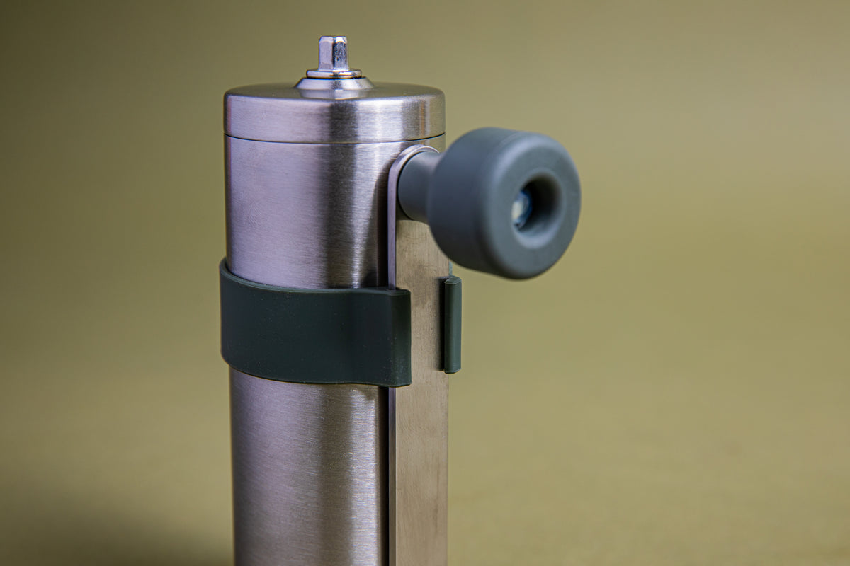 Close up of a stainless steel cylindrical shaped coffee mill canister with forest green silicone band holding stainless steel handle with plastic knob. Set against an earth-tone background.