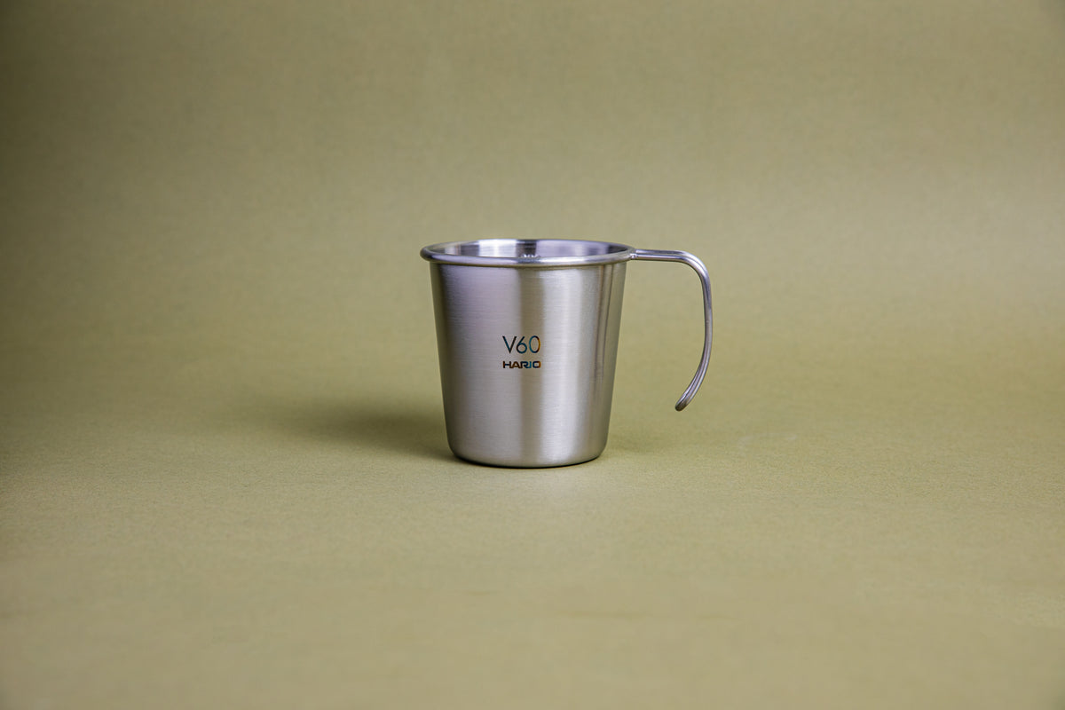 Sleek hairline silver camping mug with rounded lip and handle, etched Hario logo and V60 graphic, and set against a earth-tone background.