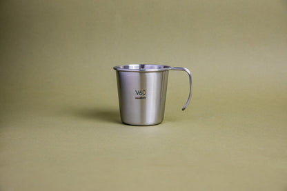 Sleek hairline silver camping mug with rounded lip and handle, etched Hario logo and V60 graphic, and set against a earth-tone background.