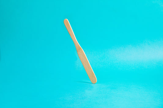 Flat, smooth, bamboo stirring stick with rounded edges, tapered between the handle and stirring portion, and set against a blue background. This item is reminiscent in shape to a tongue depressor and works great for evenly stirring coffee grounds.