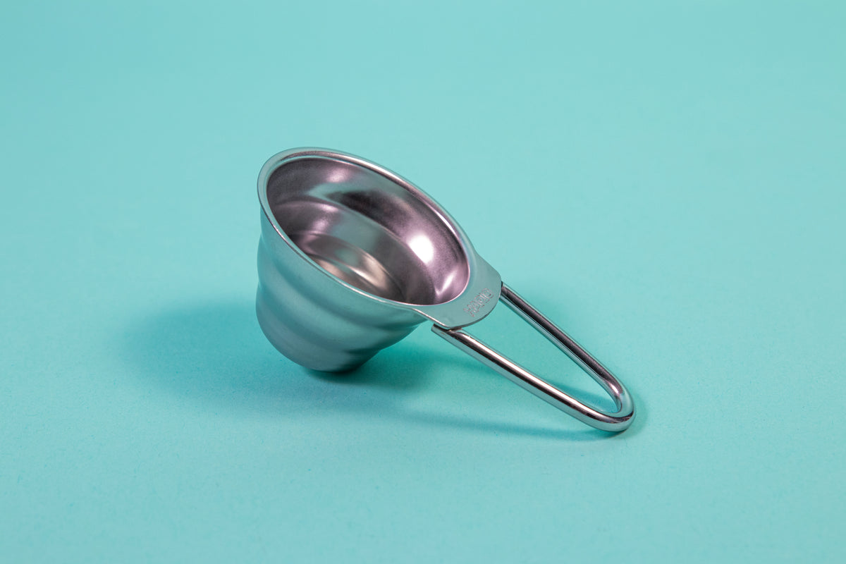 Stainless steel beehive shaped measuring spoon with Bézier shaped open air handle on a blue background.