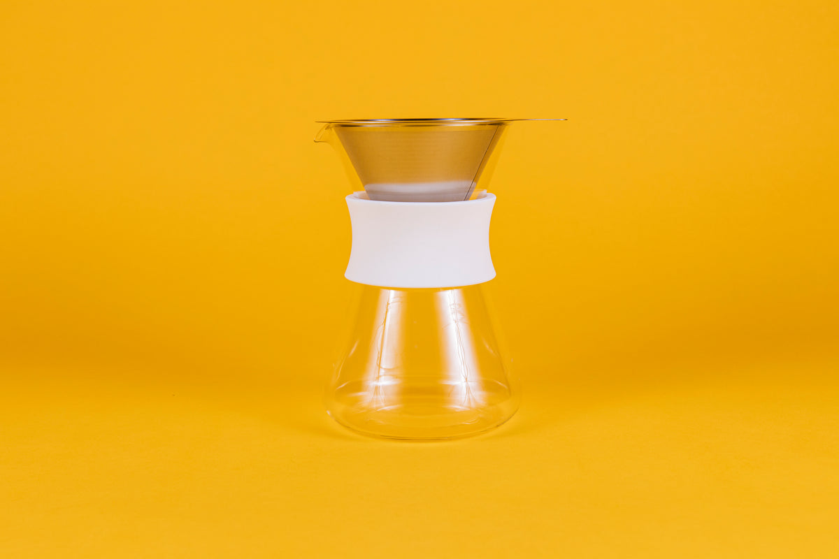 Clear glass server with white silicone collar and stainless steel mesh filter against a yellow background.