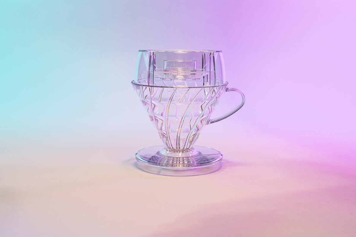 Clear plastic cone shaped dripper with clear plastic round drip assist seated on top and set against a blue and purple gradient background.