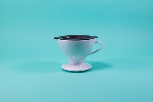 White ceramic cone shaped dripper with rounded edges and a black plastic filter insert on a blue background.