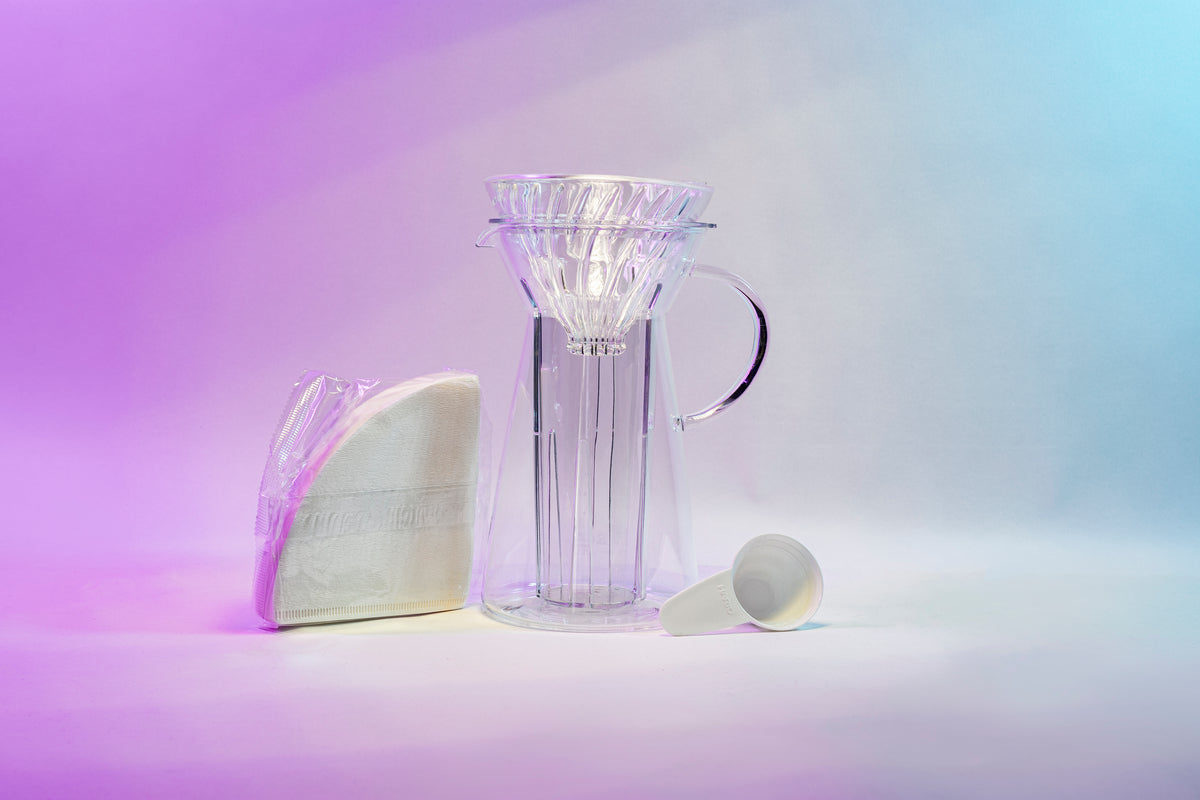 White cone shaped paper filters in a clear plastic bag next to a tapered glass dripper with fluted spout and all glass handle with a cone shaped glass dripper with ribs seated inside, and a white plastic coffee measuring spoon.