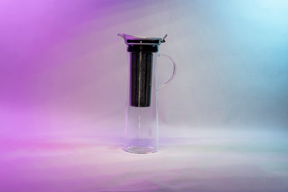 Tall, slim glass coffee pitcher with all glass full handle, stainless steel filter basket, metal lid with flared spout, and black silicone rubber seal against a purple, white, and blue background.