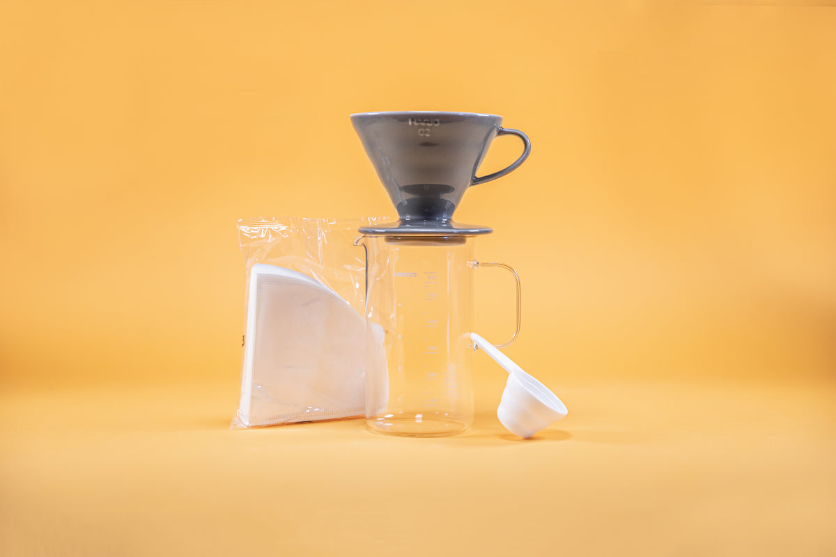 Grey ceramic coffee dripper sitting atop glass coffee server with milliliter markings from 100 to 600 and full glass handle, white paper triangular shaped coffee filters in clear plastic bag, and white plastic coffee measuring spoon against a yellow background.