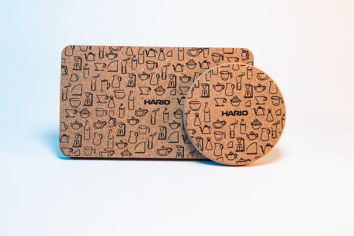Rectangular cork coaster mat with black Hario logo and black pattern design of Hario products behind a circular cork coaster with black Hario logo and black pattern design of Hario products.
