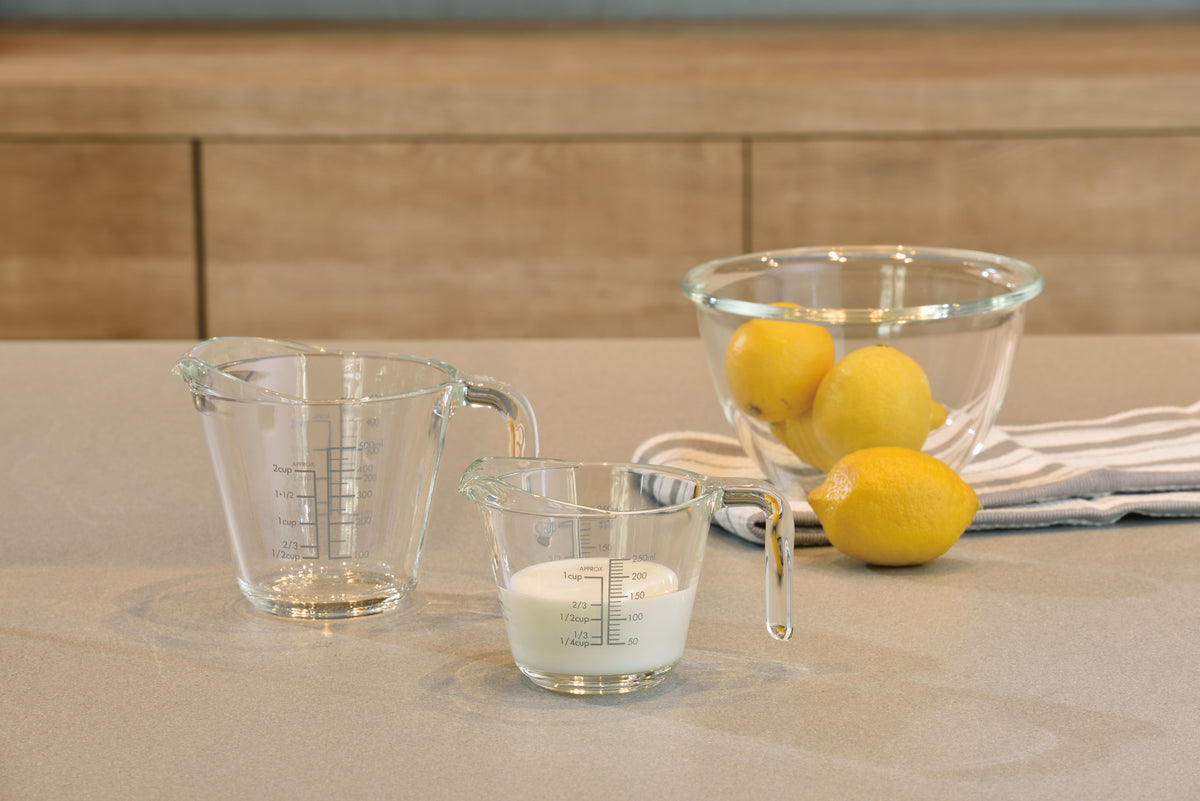One and two cup sized thick glass measuring cups with glass handles and measurements on both the outside and inside of the glass on a table next to a glass mixing bowl with lemons inside and seated on a hand towel. Beside the towel is another lemon and the one cup measuring glass is filled two-thirds of the way with milk.